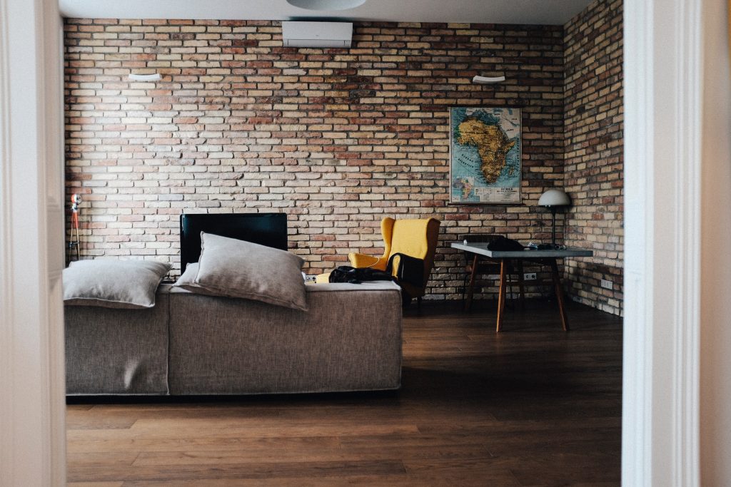 Uma sala em estilo rústico, com um grande sofá cinza, paredes de tijolinhos, um quadro de um mapa, uma mesinha com um abajur, uma cadeira amarela e uma tv