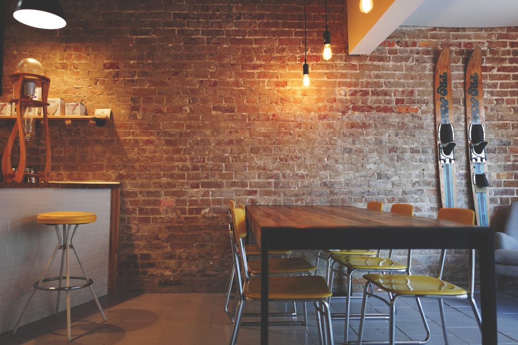 Um restaurante em estilo rústico, com as paredes de tijolos, uma mesa de madeira, bancos e cadeiras amarelos e luzes penduradas para iluminar a mesa. O balcão também é de madeira junto com uma imitação de tijolos brancos.