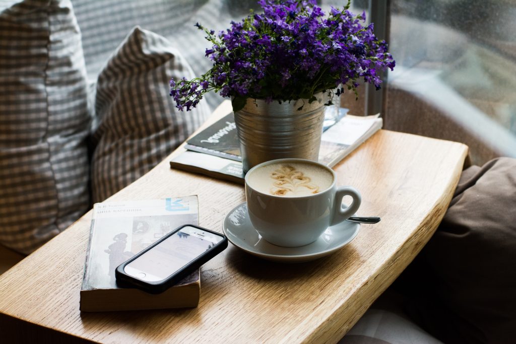 Uma bandeja em que foi aplicado acabamento para madeira, sobre uma cama ou sofá, com uma xícara de café, um vaso com flores, um livro, revistas e um celular sobre ela. Ao fundo, algumas almofadas.