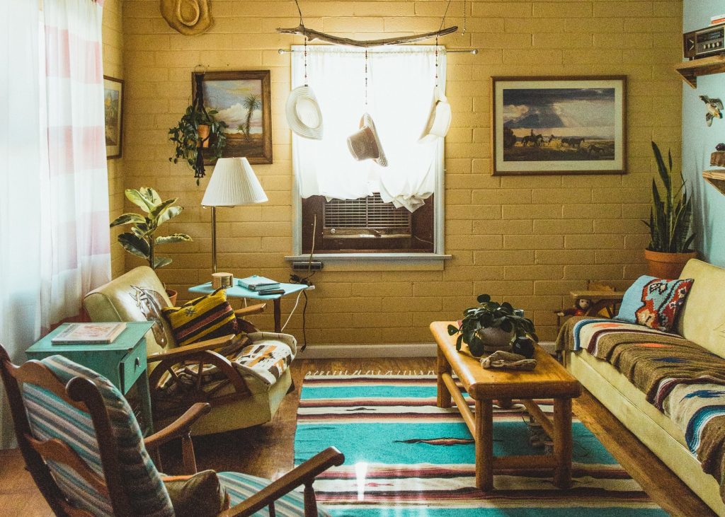 Uma sala de estilo rústico e bem aconchegante. Há um grande sofá amarelo, uma poltrona da mesma cor, uma cadeira com almofadas. A parede é de tijolos pintados de amarelo. Há quadros, chapéus e plantas pendurados. No chão de madeira, um tapete colorido e uma mesinha de madeira. No canto, há um móvel verde.