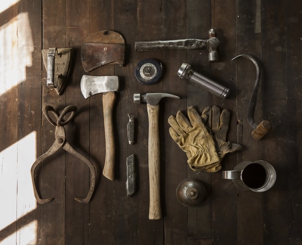 Algumas ferramentas em uma mesa de madeira, como martelo, machadinho, lanterna e luvas. Materiais bem aproveitados em uma marcenaria moderna
