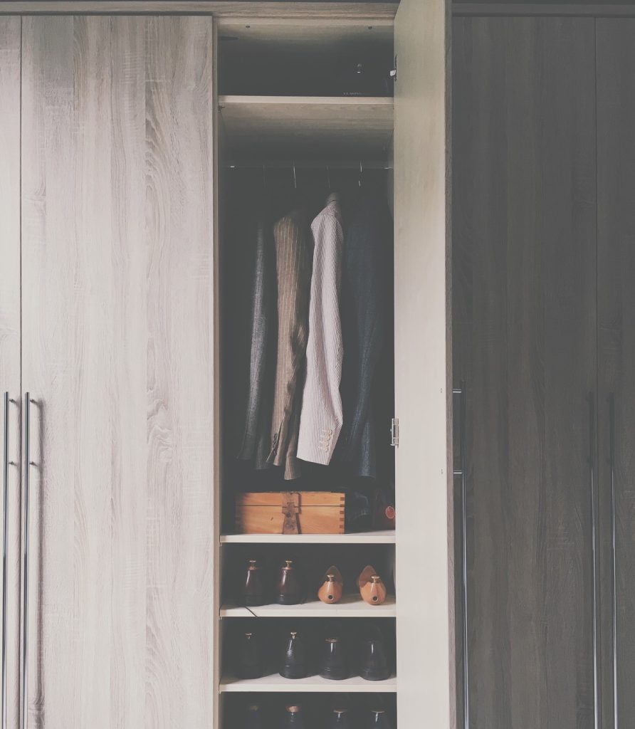 Um tipo de porta articulada de um guarda-roupas, semi-aberto. No interior, há camisas penduradas, uma caixa de madeira e sapatos nas prateleiras de baixo.