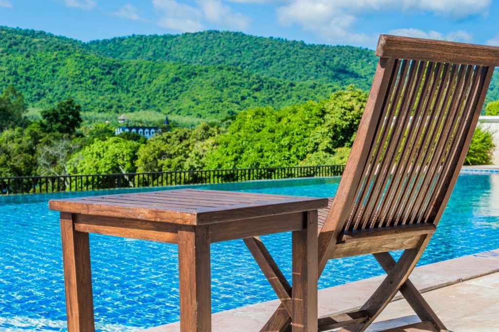 Uma mesa e uma cadeira na beira da piscina, ambas possuem acabamento para madeira adequado para o tipo de ambiente. Ao fundo, muitas árvores.