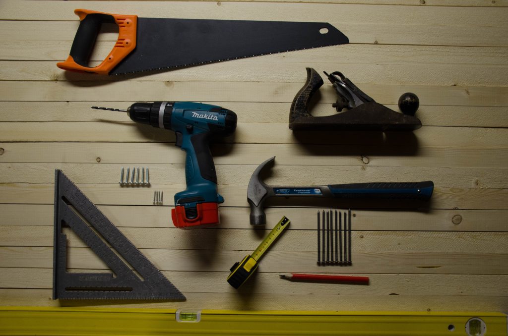 Algumas ferramentas dispostas sobre um painel de madeira, entre elas, um tipo de furadeira, um martelo, uma trena, um esquadro e um serrote