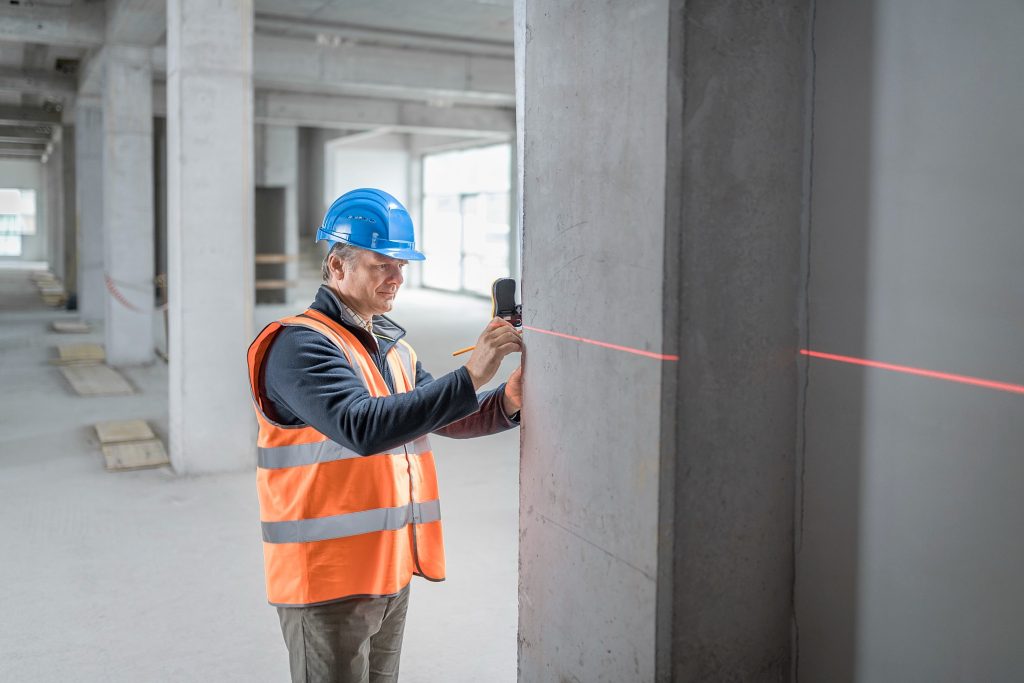 Homem branco de meia idade usando um capacete azul e colete laranja, utiliza um medidor de nível a laser em uma obra.